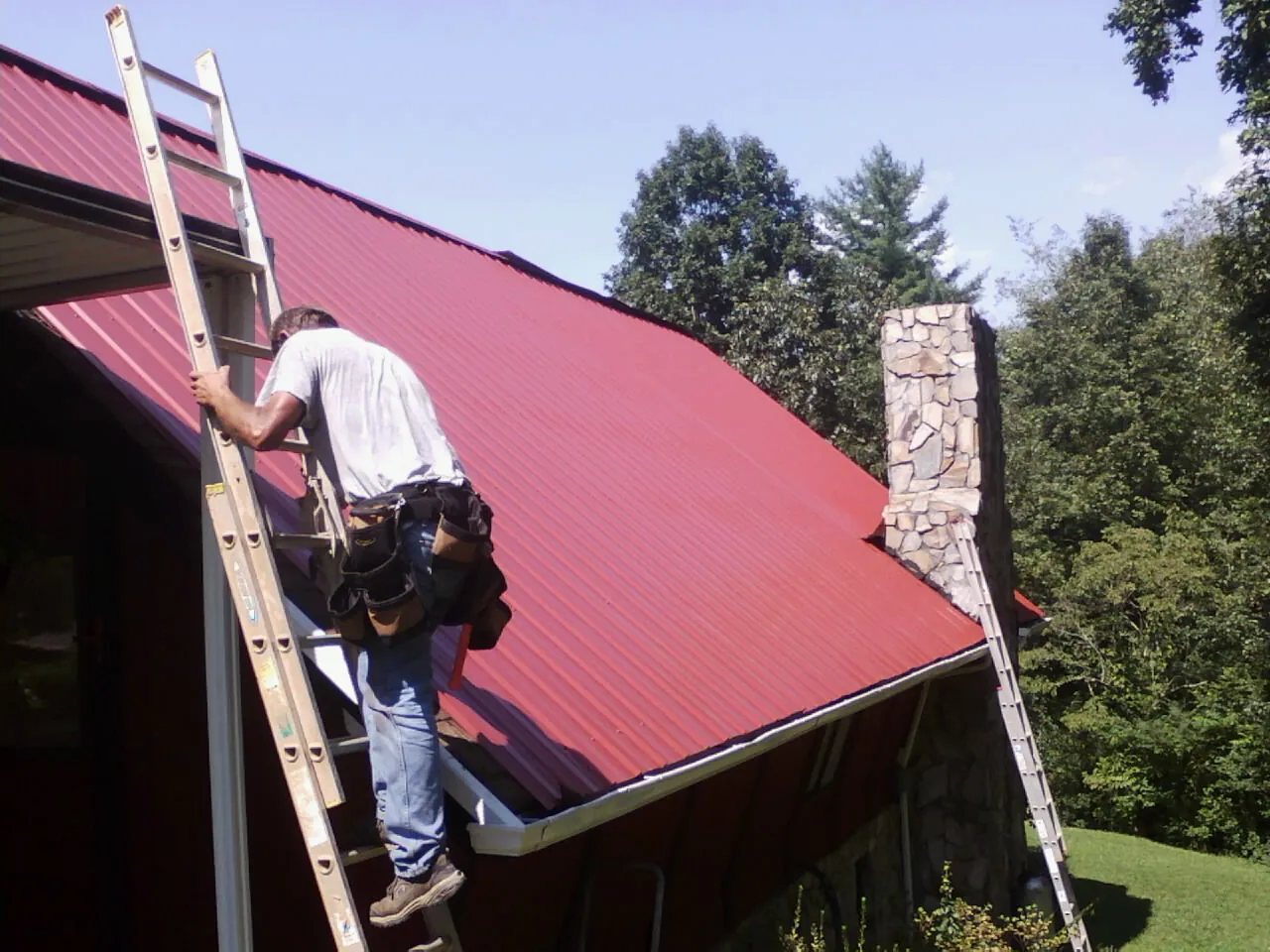 Roofer on a ladder beside a red metal roof and stone chimney — Soffit & Fascia Repair by Roofing Brothers in Obion
